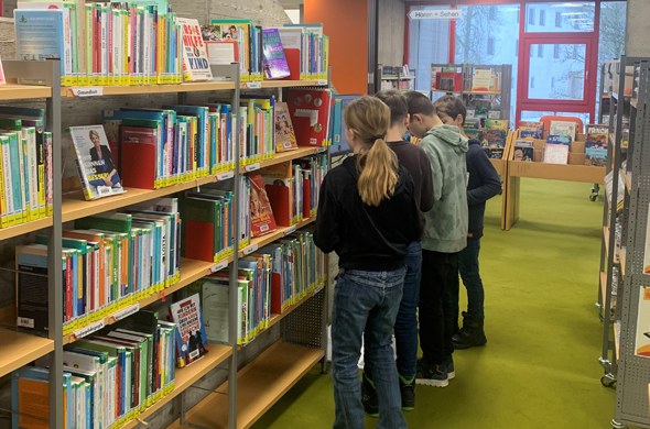 Vier Schülerinnen und Schüler stehen in Gang mit Bücherregalen und betrachten ein Buch