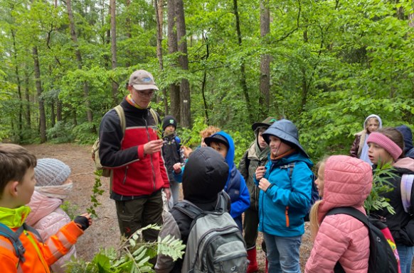 Foerster_steht_im_Wald_umringt_von_Schuelergruppe_und_erklärt_Pflanzen