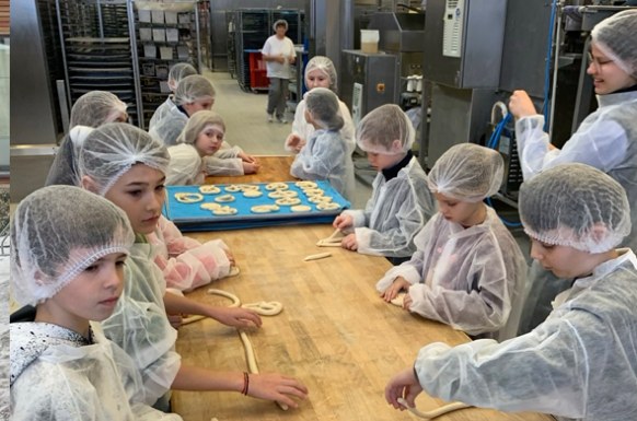 Schulkinder in der Backstube stehen an großem Backtisch und lernen das drehen von Brezen
